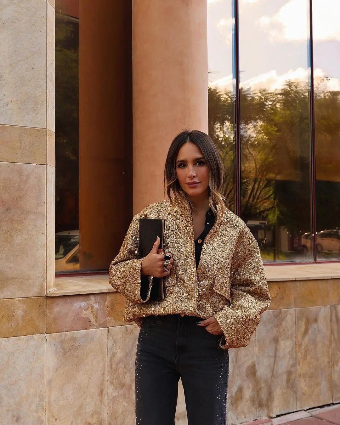 Femme habillée avec la veste bomber courte Aurélie en tissu doré à sequins, associée à un pantalon noir et une pochette élégante, un blouson chic et moderne pour un style féminin raffiné.