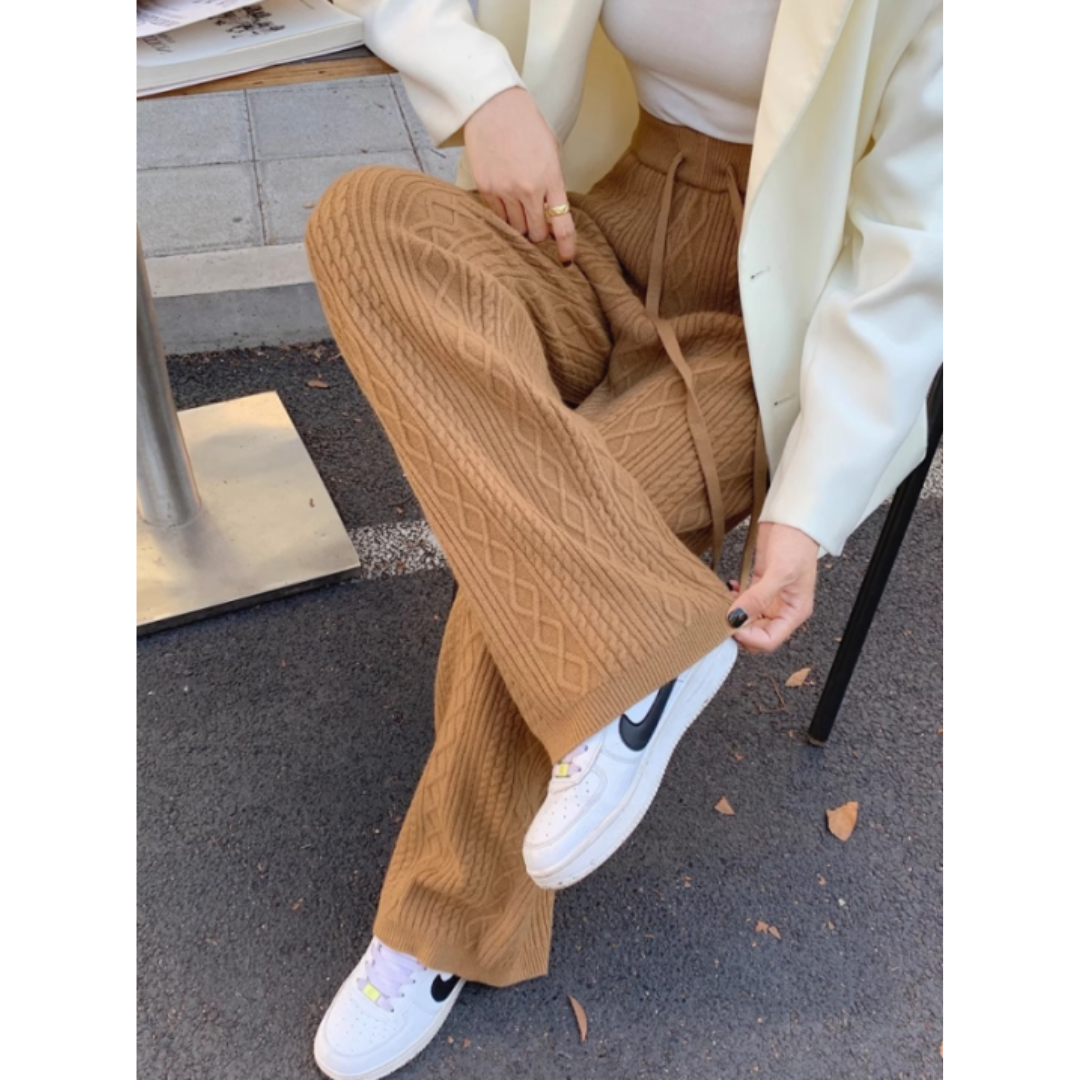 Un pantalon de jogging beige à motif en tricot, avec des jambes évasées et un cordon de serrage à la taille. Il est porté avec une paire de baskets blanches et un haut simple, offrant un look décontracté et élégant.
