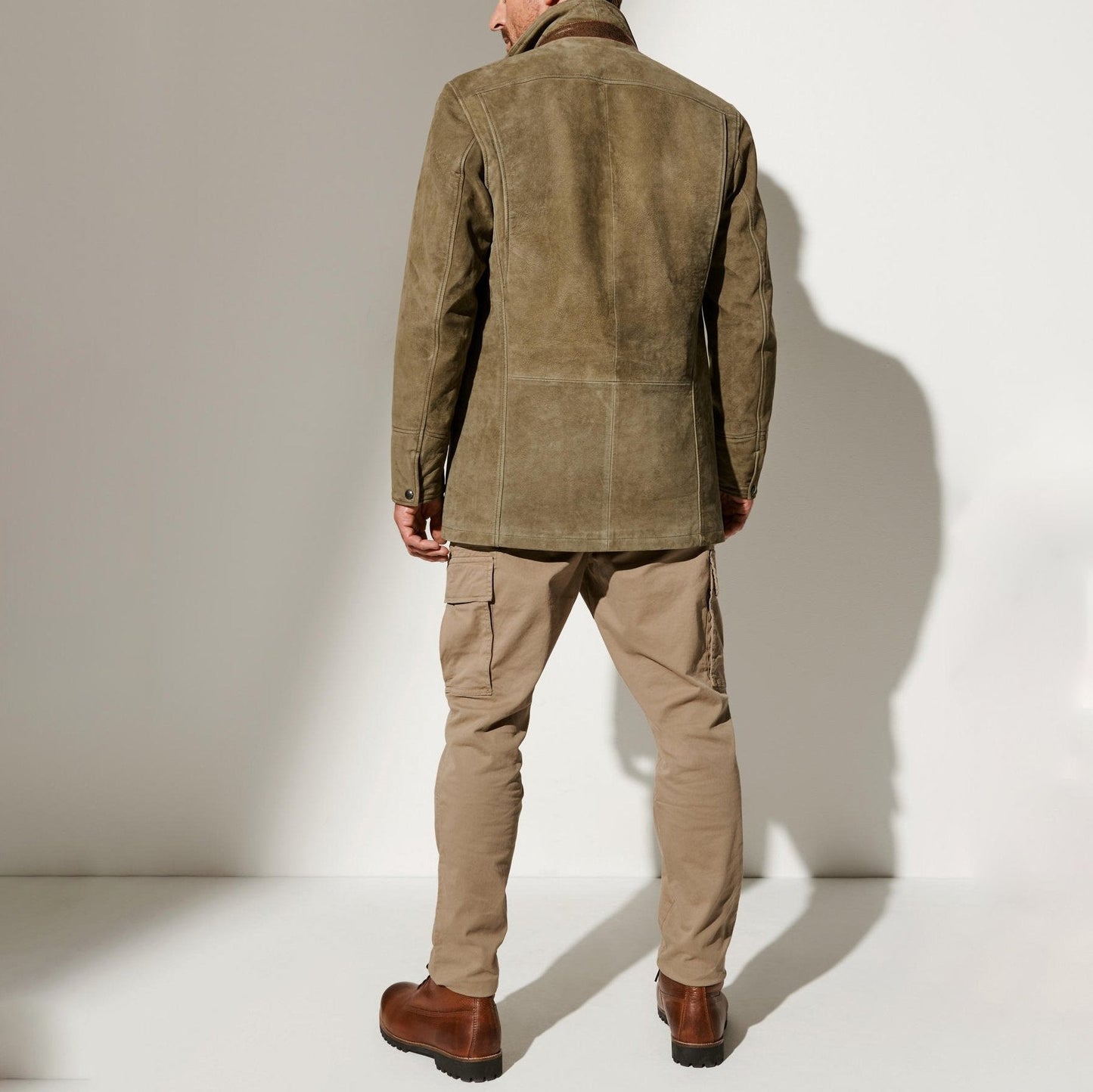Un homme est vu de dos, portant une veste en daim marron clair au style vintage, associée à un pantalon cargo beige et des bottines en cuir marron. L’ensemble crée un look masculin et sophistiqué, parfait pour une allure décontractée mais élégante. La texture de la veste et la coupe ajustée soulignent un goût raffiné pour la mode classique.