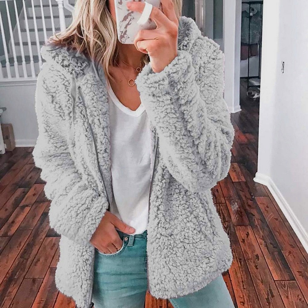 Femme portant la veste d’hiver courte Marie en fausse fourrure chaude, couleur gris clair, associée à un t-shirt blanc et un jean, pour un style cosy et tendance.