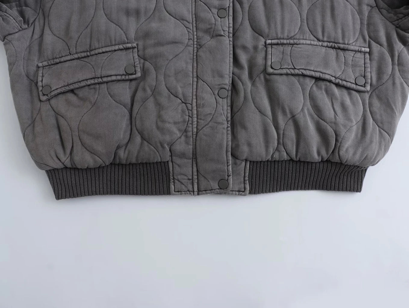 Détail du bas du blouson long pour femme "Nancy" en gris, avec un design matelassé, des poches à rabat et une finition côtelée au niveau de la taille. Parfait pour un look à la fois décontracté et élégant.
