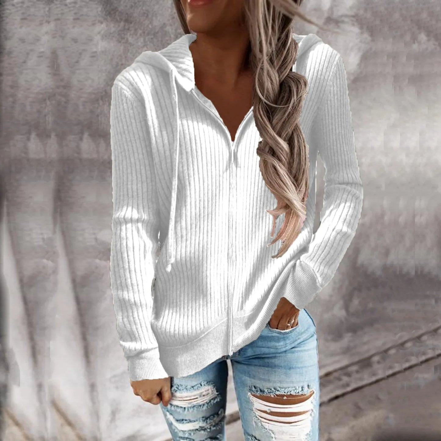 Veste à capuche zippée en maille côtelée, de couleur blanche, portée par une femme avec des cheveux longs. Elle porte un jean déchiré et affiche un look décontracté, idéal pour les journées froides.