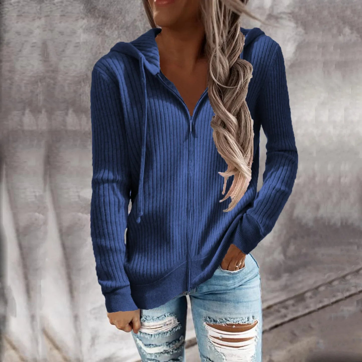 Veste à capuche zippée en maille côtelée, coloris bleu, portée par une femme souriante. Elle porte un jean déchiré et une tenue décontractée, idéale pour les journées fraîches d'automne et d'hiver.