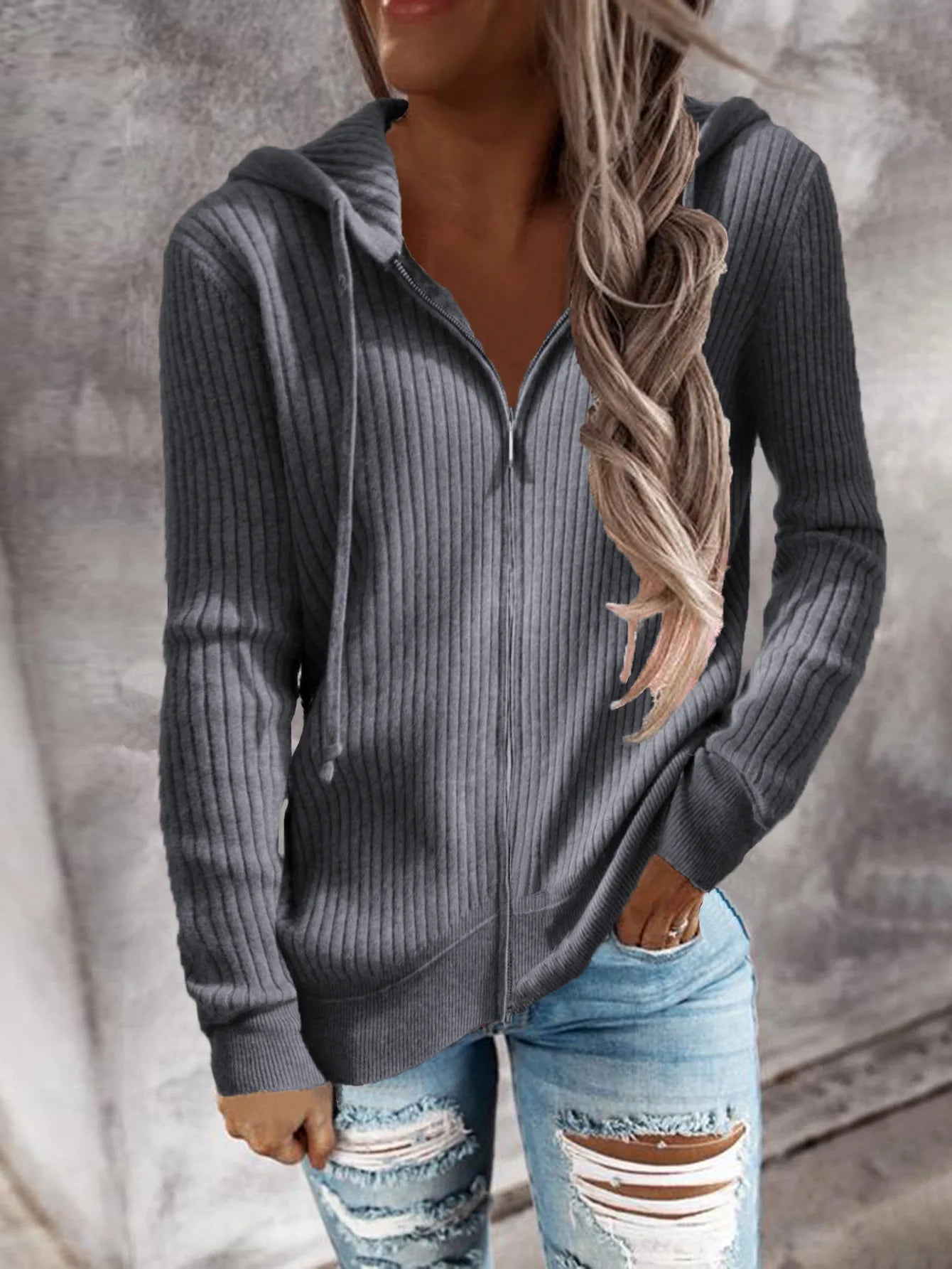 Veste à capuche zippée en maille côtelée, coloris gris, portée par une femme avec un sourire. Elle porte un jean déchiré et une coupe décontractée, idéale pour les journées fraîches d'automne et d'hiver.