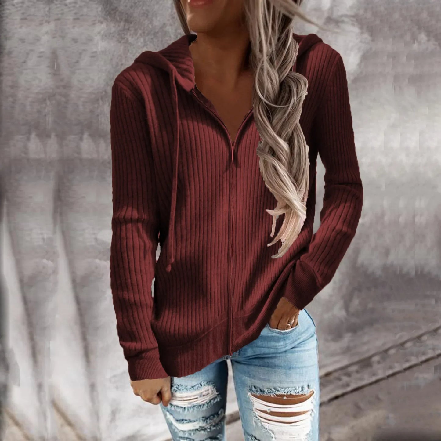 Une femme portant un sweat à capuche zippé de couleur bordeaux. Le sweat présente un motif côtelé, une coupe ajustée, des manches longues et un zip sur le devant. Elle l’associe avec un jean déchiré pour un look décontracté et moderne.