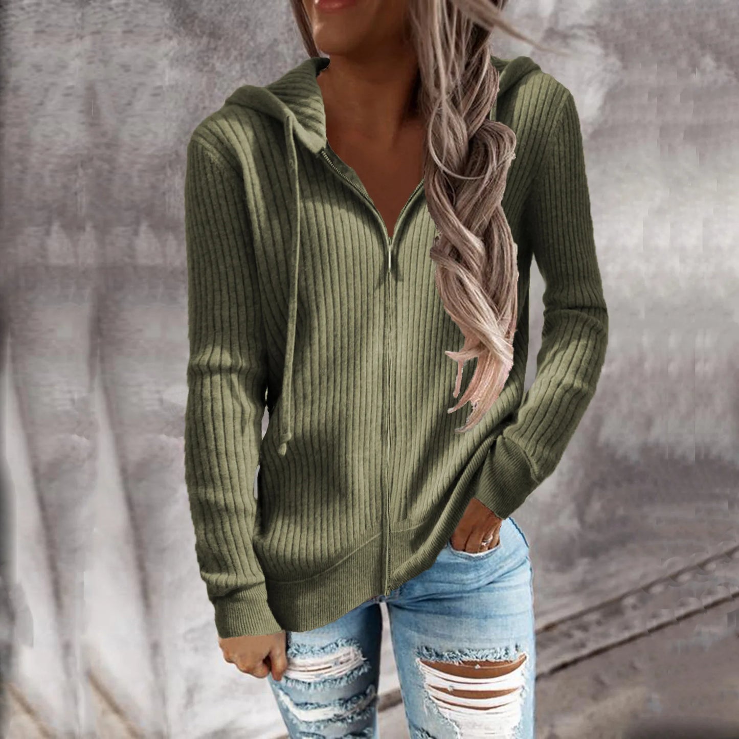 Une femme portant un sweat à capuche zippé de couleur olive. Le sweat présente un motif côtelé et une coupe ajustée, avec des manches longues et un zip sur le devant. Elle le porte avec un jean déchiré pour un look décontracté et stylé.