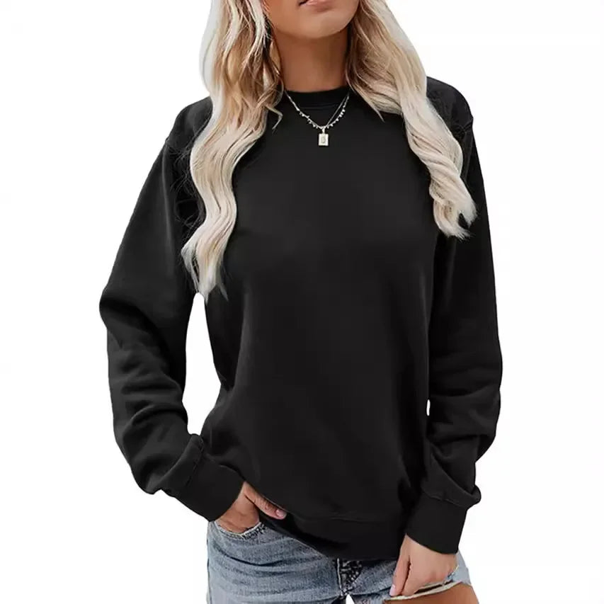 Une femme porte un sweat-shirt noir à manches longues et col rond. Elle a les cheveux blonds et porte un collier délicat, avec un jean court et déchiré, et elle pose une main sur sa taille.