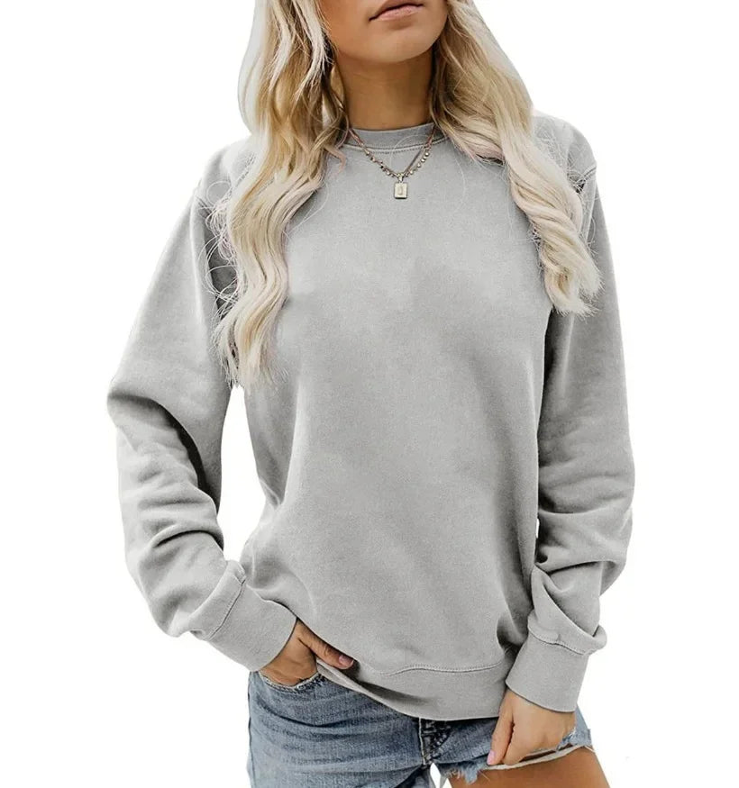 Femme portant un sweat-shirt gris clair à manches longues avec un col rond. Elle a des cheveux blonds, porte un collier délicat et un short en jean, avec une main posée sur sa taille.