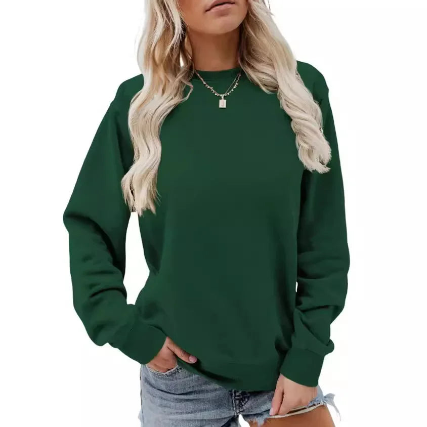 Femme portant un sweat-shirt vert foncé à manches longues avec un col rond. Elle a des cheveux blonds, porte un collier délicat et un short en jean, adoptant une pose décontractée avec ses mains sur les hanches.