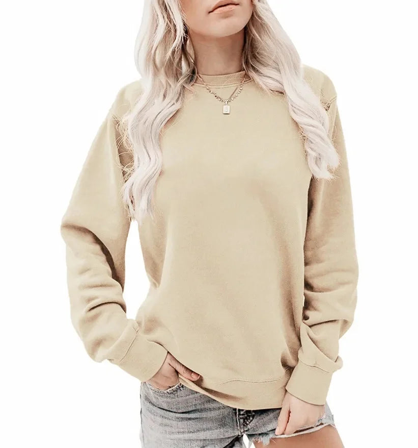 Femme portant un sweat-shirt beige à manches longues avec un col rond. Elle a des cheveux blonds, porte un collier fin et un short en jean, en affichant une pose décontractée avec une main dans sa poche.