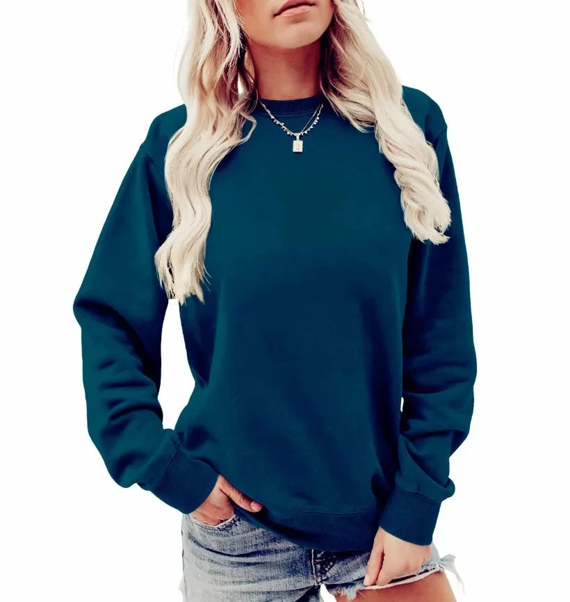 Femme portant un sweat-shirt vert foncé à manches longues avec un col rond. Elle porte un collier délicat et un short en jean, affichant une pose décontractée.