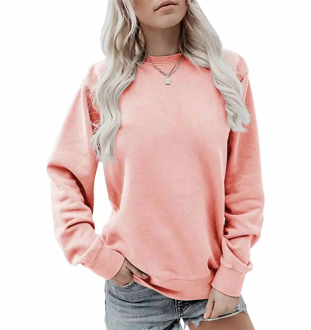 Femme portant un sweat-shirt rose à manches longues et un col rond, associé à un short en jean. Elle porte également un collier délicat et adopte une pose décontractée.