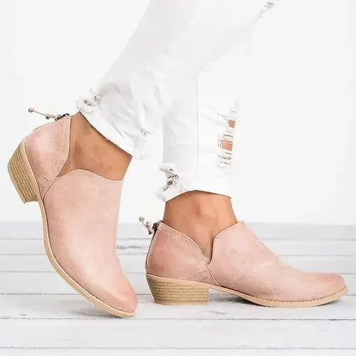 Bottines pour femme en suédine rose poudré, à talon bas épais, avec fermeture zippée à l’arrière et bout arrondi, portées avec un pantalon blanc pour un style moderne et élégant.