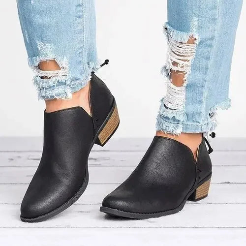 Bottines pour femme en cuir noir mat, à talon bas épais effet bois, avec bout arrondi et fermeture discrète à l’arrière, portées avec un jean bleu déchiré pour un style moderne et décontracté.