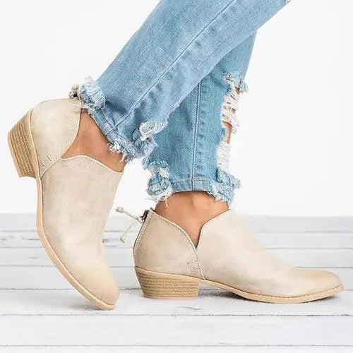 Bottines pour femme en suédine beige clair, à talon bas épais effet bois, avec bout arrondi et fermeture zippée à l’arrière, portées avec un jean bleu pour un style moderne et polyvalent.