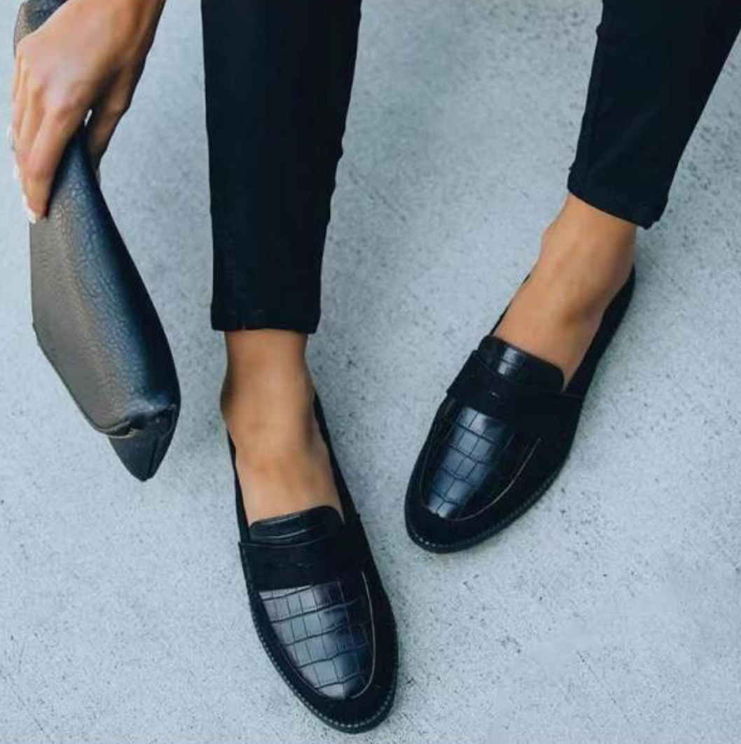 Chaussures d’été pour femme de style mocassin en cuir noir à effet texturé, avec bout arrondi et semelle plate confortable, portées avec un pantalon noir dans un décor urbain clair.