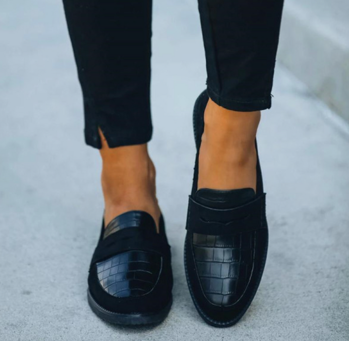 Chaussures d’été pour femme de style mocassin en cuir noir à finition texturée, avec bout effilé et semelle plate confortable, portées avec un pantalon noir dans un cadre urbain clair.