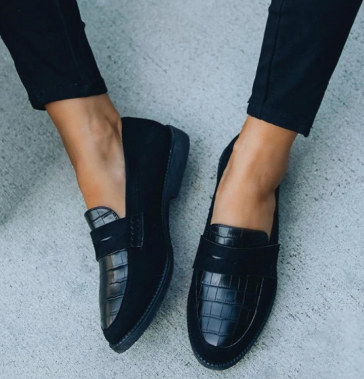 Chaussures d’été pour femme de style mocassin en cuir noir à effet crocodile, avec bride sur le dessus, bout arrondi et semelle plate confortable, portées avec un pantalon noir sur un sol clair.