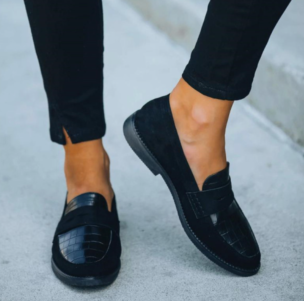 Chaussures d’été pour femme de style mocassin en cuir noir à finition texturée, avec bride sur le dessus, bout arrondi et semelle plate antidérapante, portées avec un pantalon noir en extérieur.