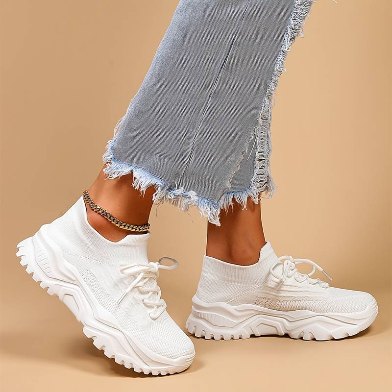 Modernes, baskets blanches à lacets et semelle épaisse crantée, portées avec un jean gris effiloché sur fond beige.