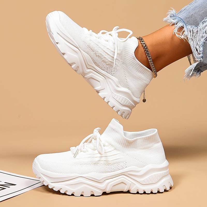 Modernes, baskets blanches à lacets en maille avec semelle épaisse crantée, une paire au sol et l’autre portée en l’air sur fond beige.