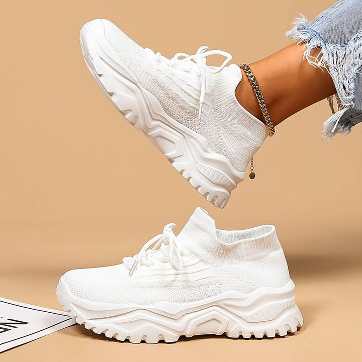 Modernes, baskets blanches à lacets en maille avec semelle épaisse crantée, une paire au sol et l’autre portée en l’air sur fond beige.