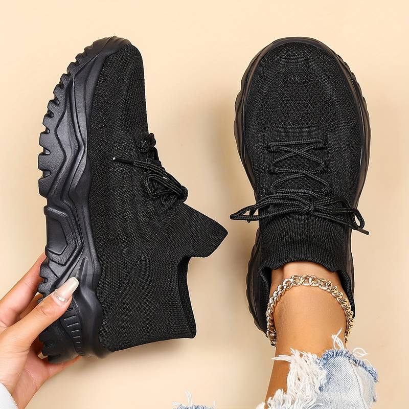 Modernes, baskets noires en maille à lacets avec semelle épaisse, une chaussure tenue à la main et l’autre portée, sur fond beige.