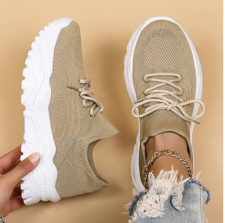 Modernes, baskets kaki/beige en maille à lacets avec semelle blanche épaisse, une chaussure tenue à la main et l’autre portée, sur fond beige.