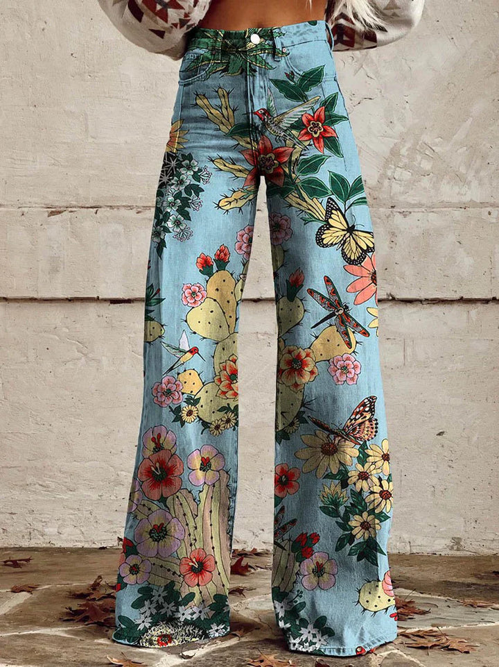 Jean ample pour femme Anouk taille haute, denim bleu clair à imprimé jardin avec fleurs, cactus et papillons multicolores, coupe large et jambes évasées, porté devant un mur en béton.