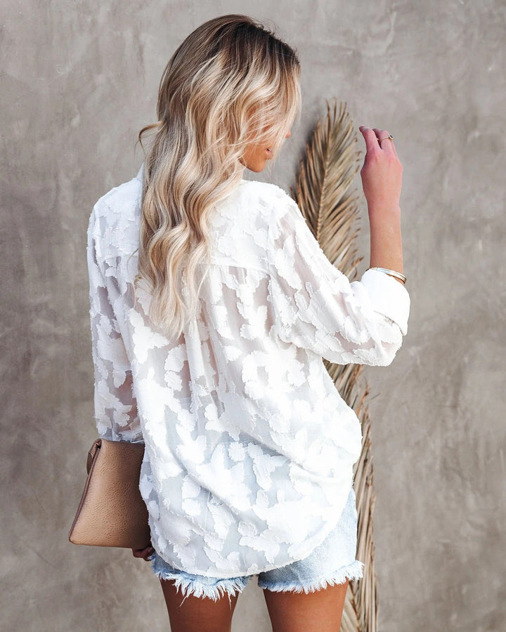 Vue arrière d’un chemisier blanc à motifs texturés pour femme, avec coupe moderne et fluide, manches longues retroussées et tissu léger semi-transparent, porté avec un short en jean pour un style chic et décontracté.