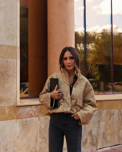 Femme habillée avec la veste bomber courte Aurélie en tissu doré à sequins, associée à un pantalon noir et une pochette élégante, un blouson chic et moderne pour un style féminin raffiné.
