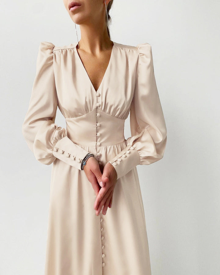 Femme élégante portant une robe en satin beige avec des manches bouffantes et des détails de boutons sur le devant, accessoirisée avec de délicats bracelets et une paire de sandales à talons hauts. La robe a une coupe sophistiquée et flatteuse, parfaite pour un événement en soirée.