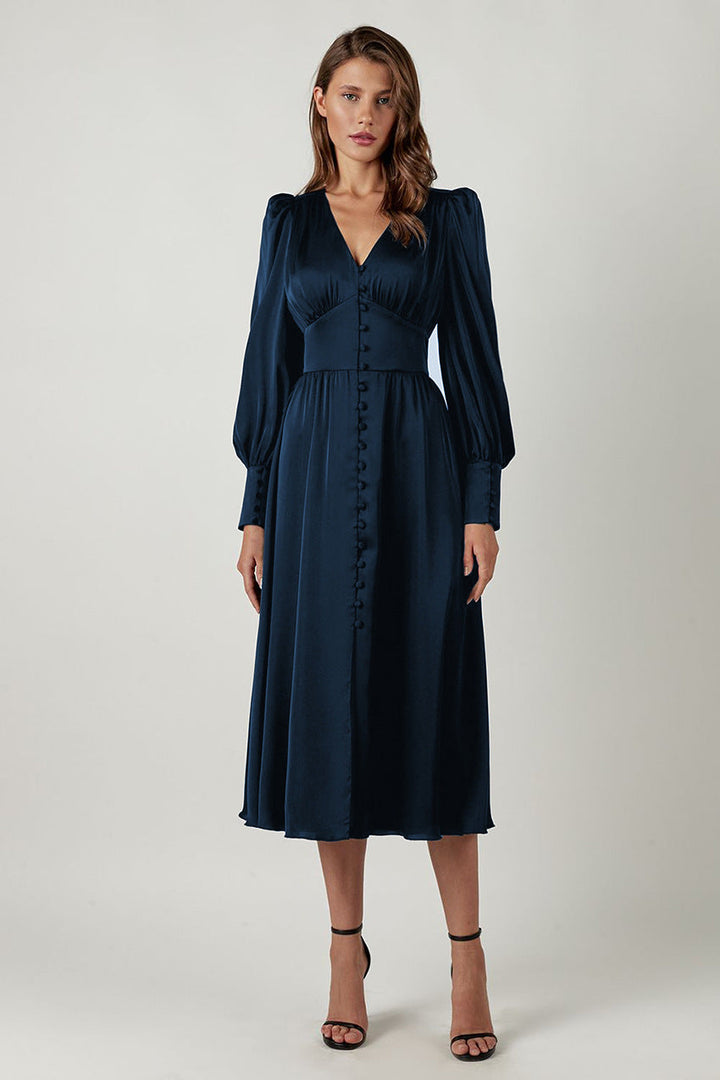 Une femme portant une robe en satin bleu marine profond, chic, avec des manches bouffantes et des détails de boutons sur le devant, associée à des talons noirs à lanières. La robe présente une coupe flatteuse avec un décolleté en V, ce qui en fait un choix parfait pour un événement élégant en soirée.