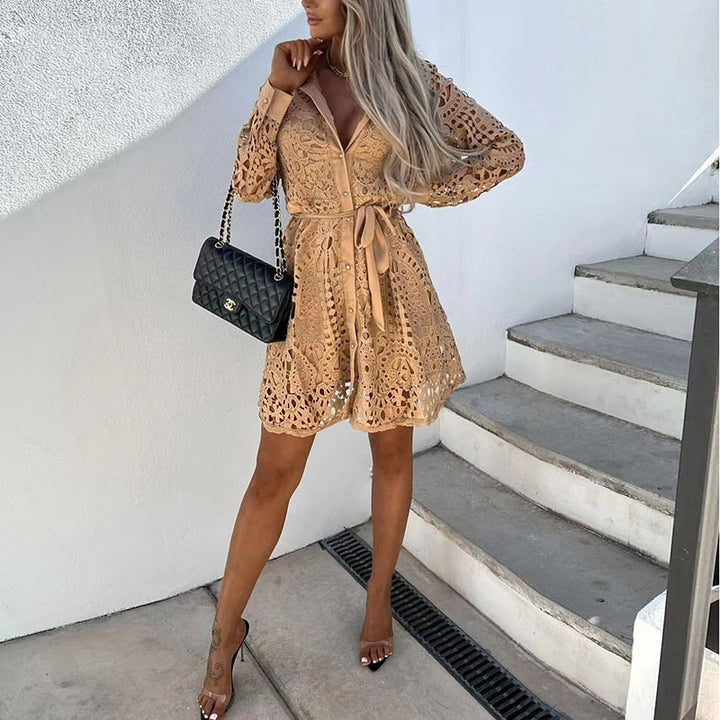 Une femme portant une robe mini en dentelle beige, au design élégant et moderne, avec des boutons sur le devant et une ceinture assortie à la taille. Elle tient un sac à main noir de marque Chanel et porte des sandales à talons transparents. La robe met en valeur un style chic et raffiné, idéal pour les occasions estivales.