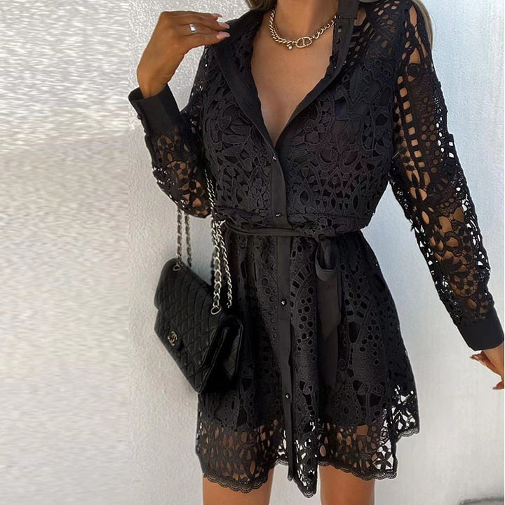 Une femme portant une robe mini noire en dentelle, avec des manches longues et une ceinture satinée qui marque la taille. Elle porte un collier en chaîne et tient un sac à main noir de marque Chanel. Cette robe élégante et moderne est parfaite pour un look sophistiqué et féminin, idéal pour des occasions spéciales.