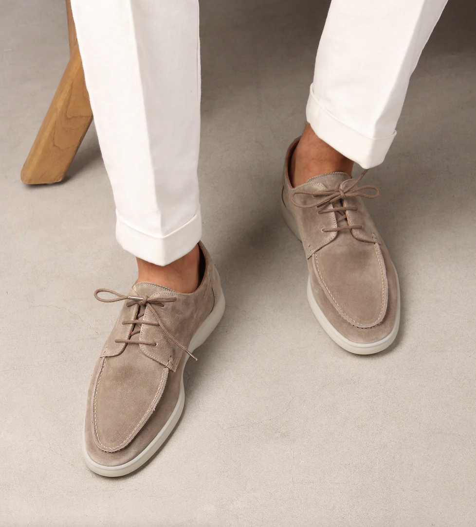 Mocassins classiques pour hommes Boniface en daim beige, à lacets et semelle claire, portés avec un pantalon blanc sur fond gris.