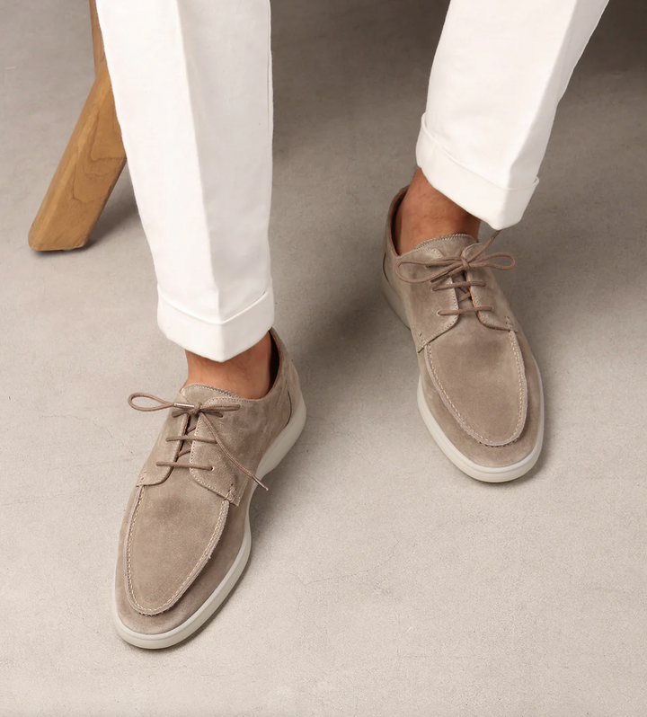 Mocassins classiques pour hommes Boniface en daim beige, à lacets et semelle claire, portés avec un pantalon blanc sur fond gris.