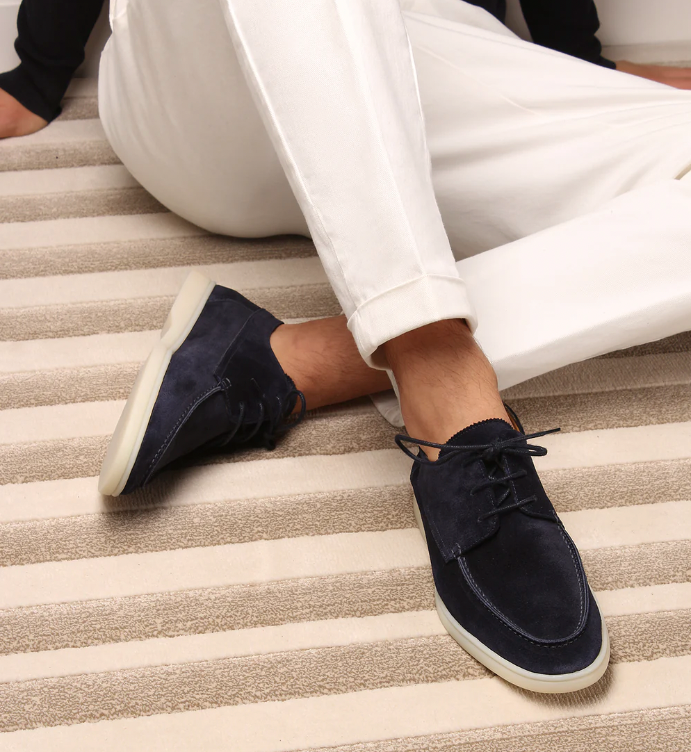 Mocassins classiques tendance pour homme en daim bleu marine, avec lacets et semelle claire, portés avec un pantalon blanc sur un tapis à rayures.