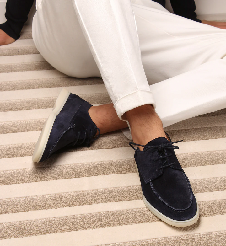 Mocassins classiques tendance pour homme en daim bleu marine, avec lacets et semelle claire, portés avec un pantalon blanc sur un tapis à rayures.