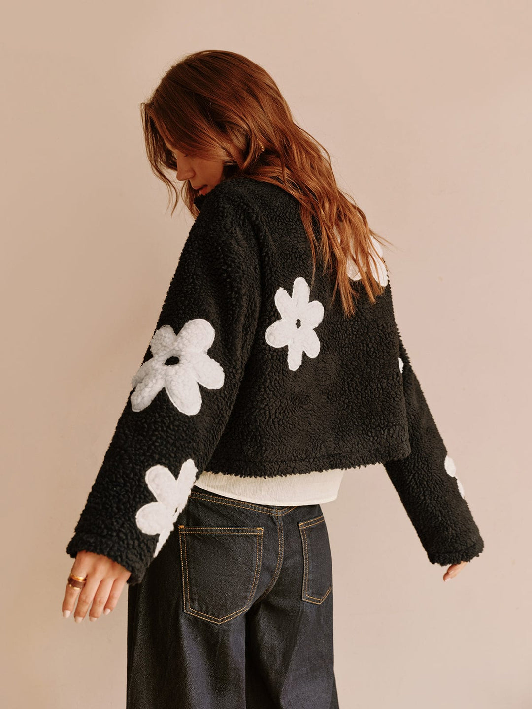 Vue arrière de cette veste courte en fausse fourrure noire ornée de fleurs blanches. Son design unique et tendance, avec des détails soignés, offre un look moderne et décontracté, parfait pour un style urbain et féminin.