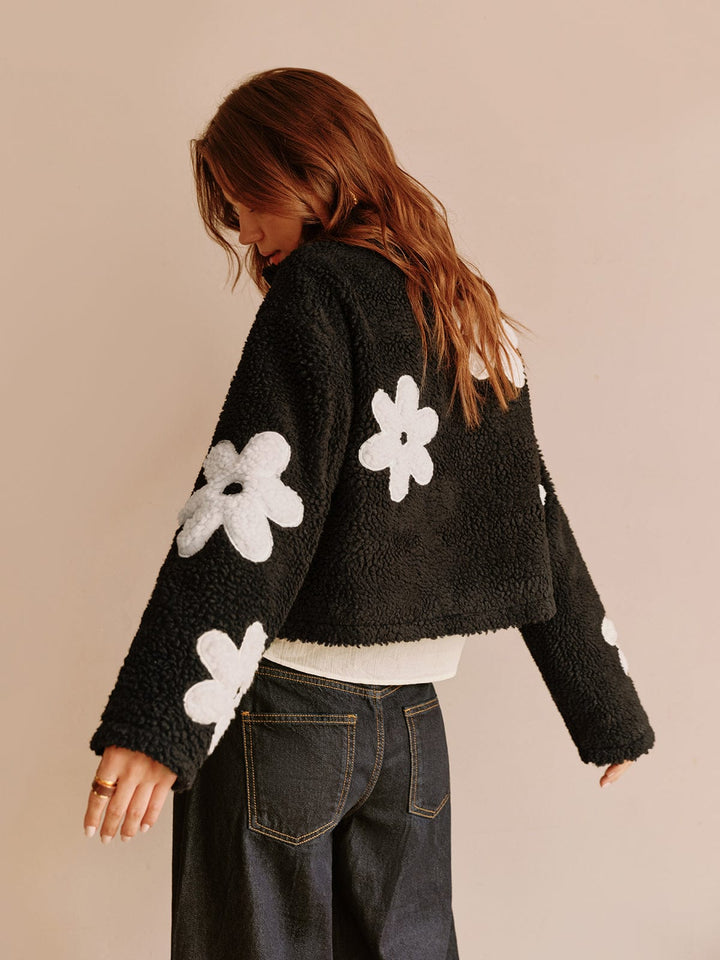 Vue arrière de cette veste courte en fausse fourrure noire ornée de fleurs blanches. Son design unique et tendance, avec des détails soignés, offre un look moderne et décontracté, parfait pour un style urbain et féminin.