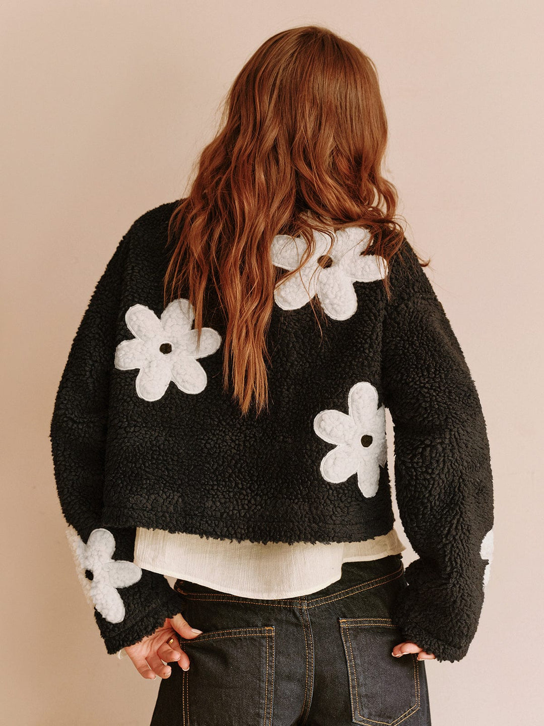 Vue arrière de cette veste courte en fausse fourrure noire avec des fleurs blanches, parfaite pour un look à la fois tendance et confortable. Associez-la à un top en tissu léger et un pantalon taille haute pour une allure moderne et féminine.