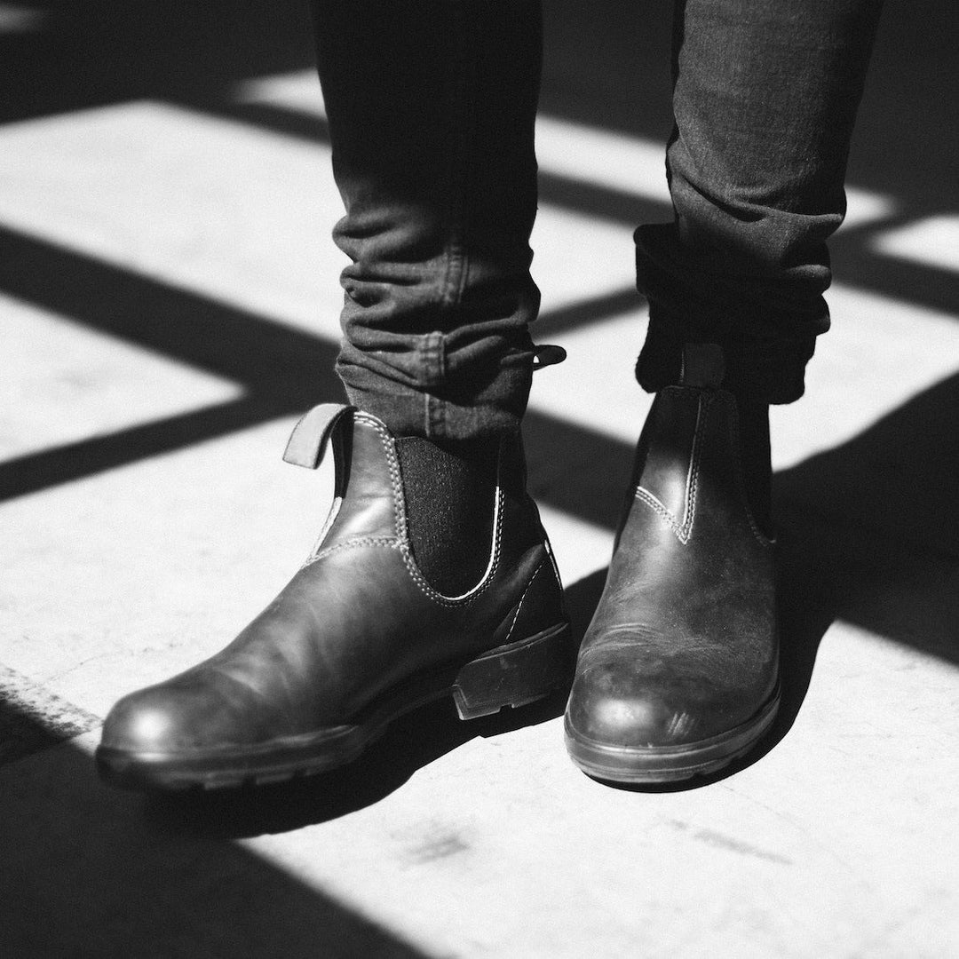 Vue rapprochée d’une paire de bottines noires en cuir portées avec un jean ajusté. Les bottines affichent une finition lisse et mate, avec des coutures apparentes et des empiècements élastiques sur les côtés pour un confort optimal. La photo, en noir et blanc, met en valeur leur style minimaliste et intemporel, parfait pour un look chic et décontracté.