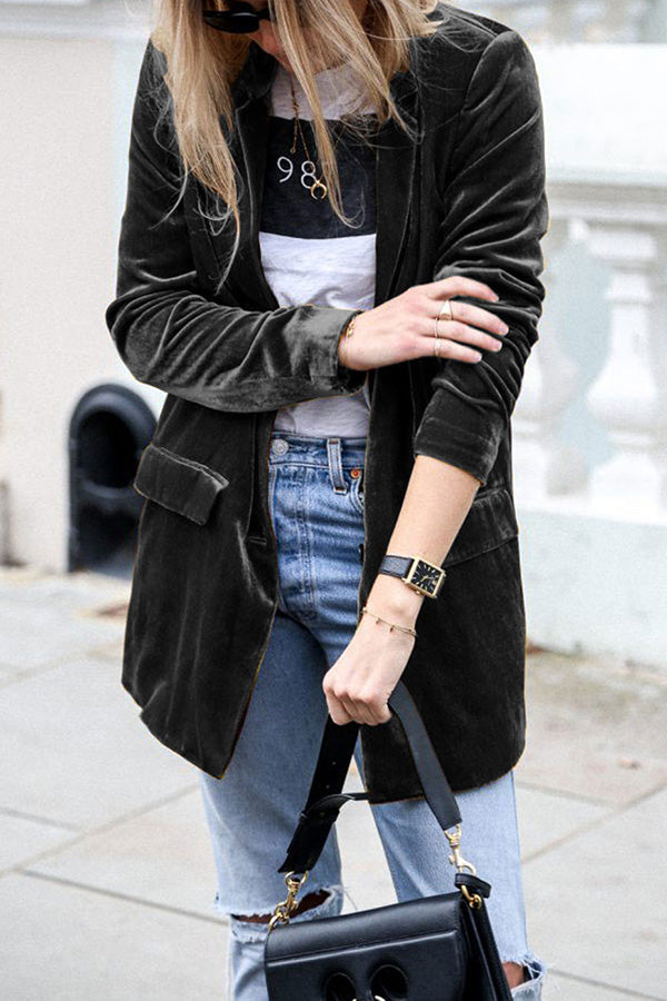 Femme portant une veste en velours gris foncé, un t-shirt blanc imprimé et un sac à main noir. Elle porte un jean clair légèrement déchiré et ajuste son sac, créant un look à la fois chic et décontracté, parfait pour un style urbain moderne.