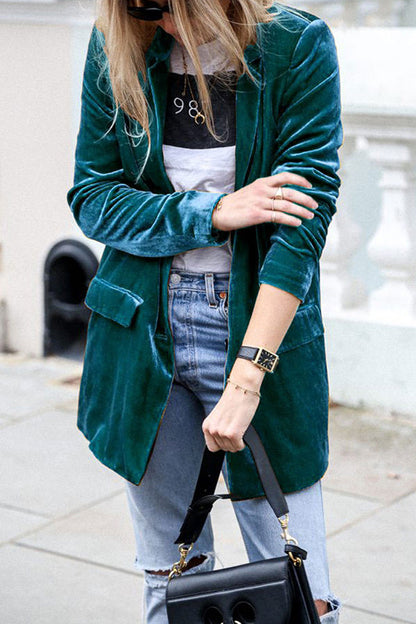 Femme portant une veste en velours vert émeraude, un t-shirt blanc imprimé et un sac à main noir. Elle porte un jean clair légèrement déchiré et ajuste son sac, créant un look élégant et moderne, parfait pour un style urbain et décontracté.