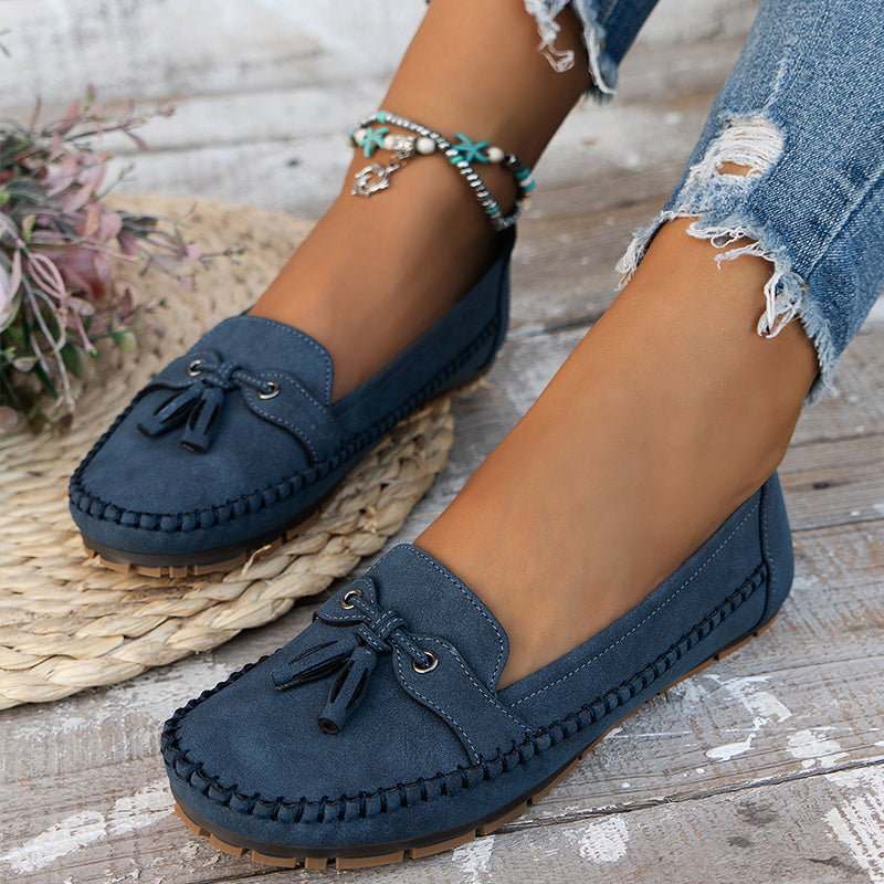 Des mocassins plats en suède bleu marine, dotés d'un nœud décoratif et d'une semelle en caoutchouc pour un confort optimal. Ce modèle, associé à un jean déchiré et des accessoires bohèmes, incarne un style décontracté et moderne, parfait pour un look chic et confortable.