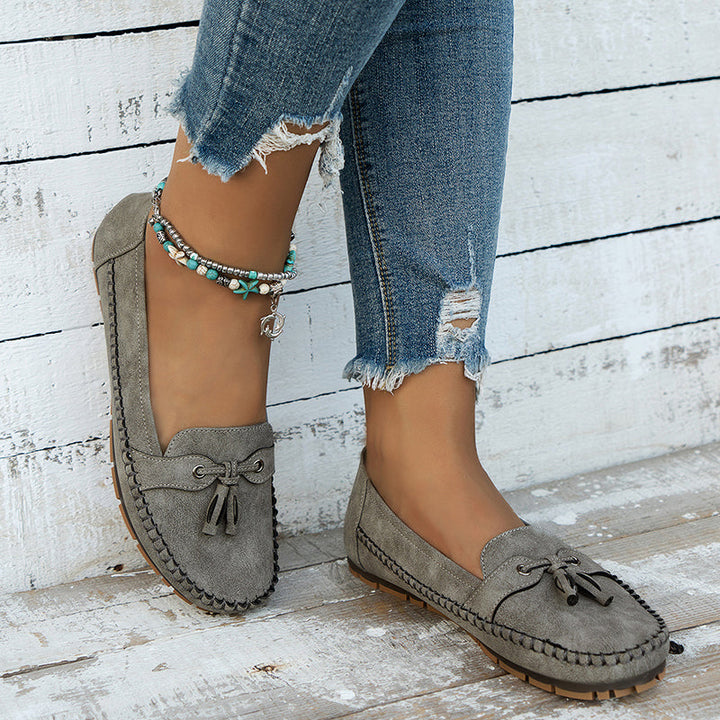 Des mocassins plats gris en suède, avec un nœud décoratif à l'avant, parfaits pour un style décontracté et moderne. Ils sont associés à un jean déchiré et un bracelet bohème, créant une tenue chic et confortable. Un choix idéal pour allier confort et mode au quotidien.