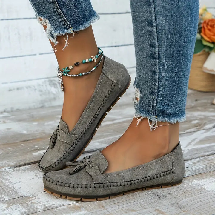 Vue latérale des mocassins plats gris pour femmes, avec un nœud décoratif et une finition en suède. Ces mocassins sont parfaits pour un look décontracté et moderne, complétés par des bracelets bohèmes et un jean déchiré. Confort et style assurés pour toutes les occasions.