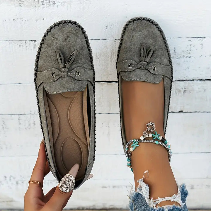 Vue de dessus des mocassins plats pour femmes, en suède gris avec un design élégant et un nœud décoratif. Le confort de la semelle intérieure et le détail du bracelet bohème sur le pied apportent une touche de style décontracté et moderne. Parfait pour un look chic et confortable.