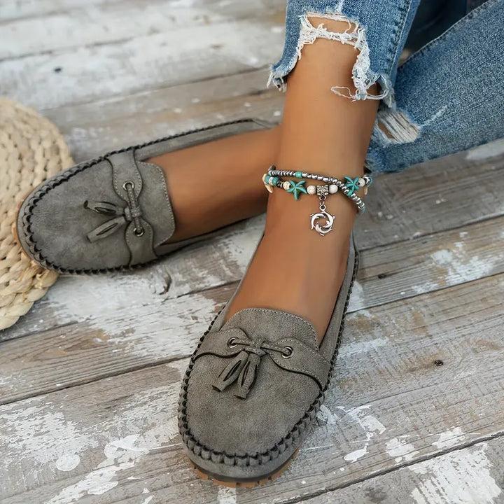 Des mocassins modernes pour femmes, en suède gris avec un nœud décoratif à l'avant. Ce modèle élégant et confortable s'associe parfaitement avec un jean déchiré et des accessoires comme un bracelet bohème. Idéals pour un look décontracté et tendance.